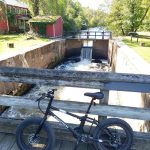 Mountain bike on wooden dock overlooking a small dam with flowing water and trees.