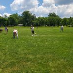 People playing a game of flag football on a sunny, grassy field surrounded by trees.