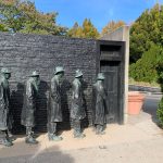 Bronze statues of workers stand before a dark, textured wall and doorway outdoors.