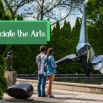 People view a modern, metallic sculpture outdoors amidst lush greenery.