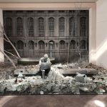 Sculpture featuring a figure amidst rubble, set against a backdrop of an old building facade.