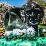 Giant bronze head sculpture with sheep figures in a blooming park setting.