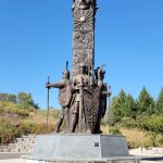 Monument featuring a warrior head atop figures in armor against a clear blue sky.