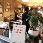 Woman and barista in cafe with "Pick Up Here" sign visible.