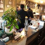Bar scene with staff preparing drinks behind a counter, featuring plants and menu boards.