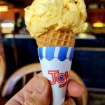Yellow ice cream in a blue and white Joy branded waffle cone held by a hand.