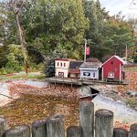 Rustic red barn and small buildings near a muddy creek surrounded by autumn trees.