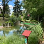 Golf course scene with a pond, lush greenery, and trees under a blue sky.