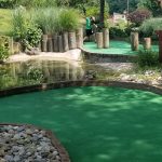 Putting green with water hazard and golfer practicing in a lush outdoor setting.