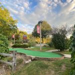 Mini golf course with green putting surface, trees, and blue sky.