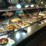 Buffet line with various food trays under overhead lighting in a dining area.