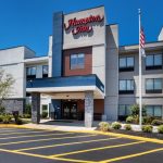 Modern Hampton Inn hotel exterior with a large parking lot and clear blue sky.
