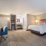 Modern hotel room with king bed, neutral decor, and a small sitting area.