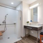 Modern hotel bathroom with white shower, vanity mirror, and wood-look flooring.