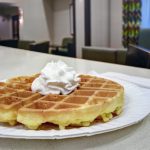Waffle topped with whipped cream on a white plate, set on a counter in a bright cafe.