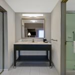 Modern hotel bathroom vanity area with mirror, sink, and adjacent glass shower enclosure.