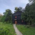 Old black wooden barn nestled in a lush, green, wooded landscape under a cloudy sky.