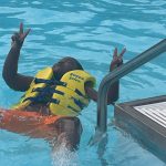Child in yellow life vest making peace signs while swimming near a pool ladder.