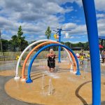Woman plays under colorful water arch structure at a sunny playground.