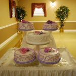 Three tiered cakes displayed on a white table in a warmly lit room.