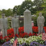 War memorial with five stone soldiers and a central obelisk surrounded by colorful flowers.