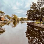 Calm river flowing under a cloudy sky, lined with trees and stone embankment.