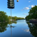 Pagoda-like lamp post over calm river reflecting blue sky and green trees.