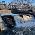 Stone weir with cascading water under an ornate metal bridge on a clear day.