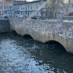 Stone arch bridge over a flowing river with buildings lining the street under a bright sky.