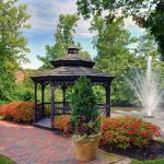 Gazebo by a pond with a fountain, surrounded by lush green trees and colorful flowers.