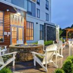 Outdoor patio area with stone fire pit, white Adirondack chairs, and modern building backdrop at dusk.