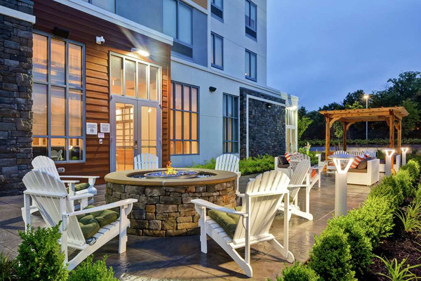 Outdoor patio area with stone fire pit, white Adirondack chairs, and modern building backdrop at dusk.