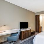 Modern hotel room with a desk, TV, and white bed.