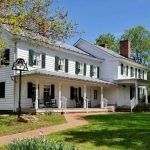 Historic white farmhouse with green shutters and porch under a bright blue sky.