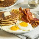 Stack of pancakes, fried eggs, and bacon on a white plate with toast nearby.