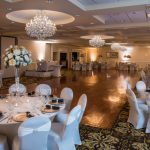 Elegant ballroom set up for an event with white tables, floral centerpieces, and crystal chandeliers.