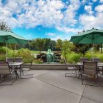 Outdoor patio with lounge chairs, green umbrellas, and a central water fountain under a blue sky.