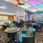 Bright hotel lounge with modern seating, colorful ceiling lighting, and a view into the kitchen area.