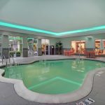 Indoor swimming pool area with bright lighting, lounge chairs, and glass-enclosed rooms.