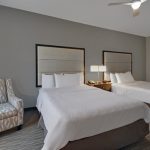 Two modern hotel beds with white linens and a textured armchair in a neutral room.
