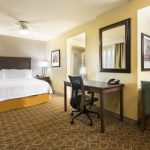 Hotel room interior showing a queen bed, desk area, and glimpse of the kitchenette.