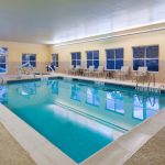 Indoor swimming pool with blue water, surrounded by lounge chairs and windows.