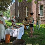 Historical reenactors in period dress gather outdoors near a stone building.