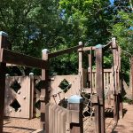 Wooden playground structure surrounded by lush green trees on a sunny day.