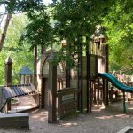 Wooden playground structure with slides nestled under leafy green trees.