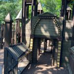 Wooden playground structure with towers and a covered play area under trees.