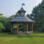 Victorian-style gazebo with a dark roof in a grassy park setting under a blue sky.