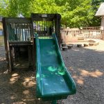 Green slide and wooden play structure in a sunny, wooded playground area.