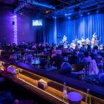Live band performance on a stage with an audience seated in a dimly lit venue.