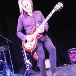 Musician with long hair plays electric guitar on a dark stage during a live performance.